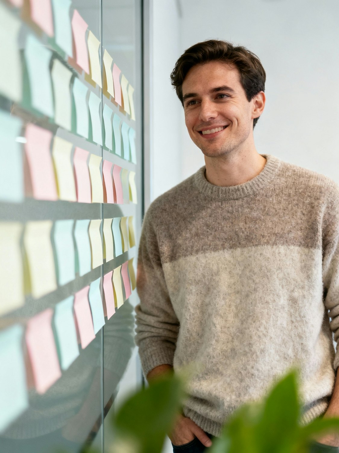A smiling teammate in a grey sweater reading rows of colored sticky notes on a glass wall.